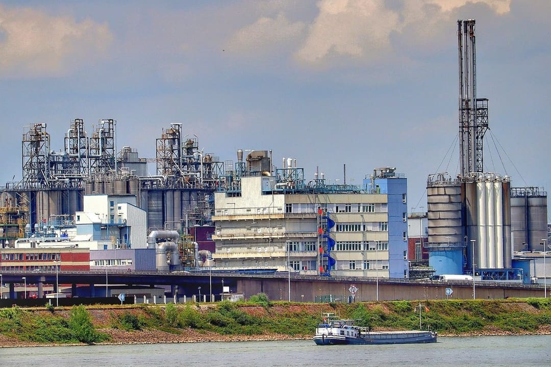 Tankowiec płynący Renem obok rafinerii z wieżami, zbiornikami i rurociągami, w tle napięć wokół dostaw ropy przez Ormuz.